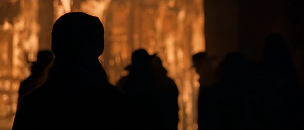 The Claim (2000) movie still: medium — Silhouettes of several figures stand before a large structure engulfed in bright orange flames. The figures…