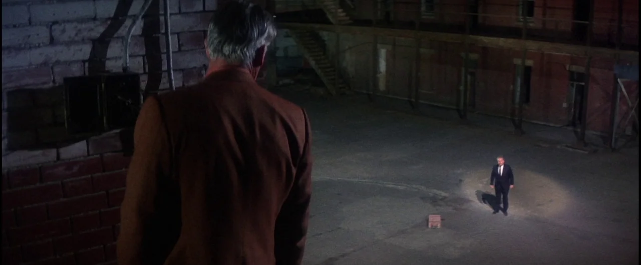 Point Blank (1967) movie still: medium — An outdoor paved space framed by a brick wall and a multi-level structure. A lone man in a suit stands in a…