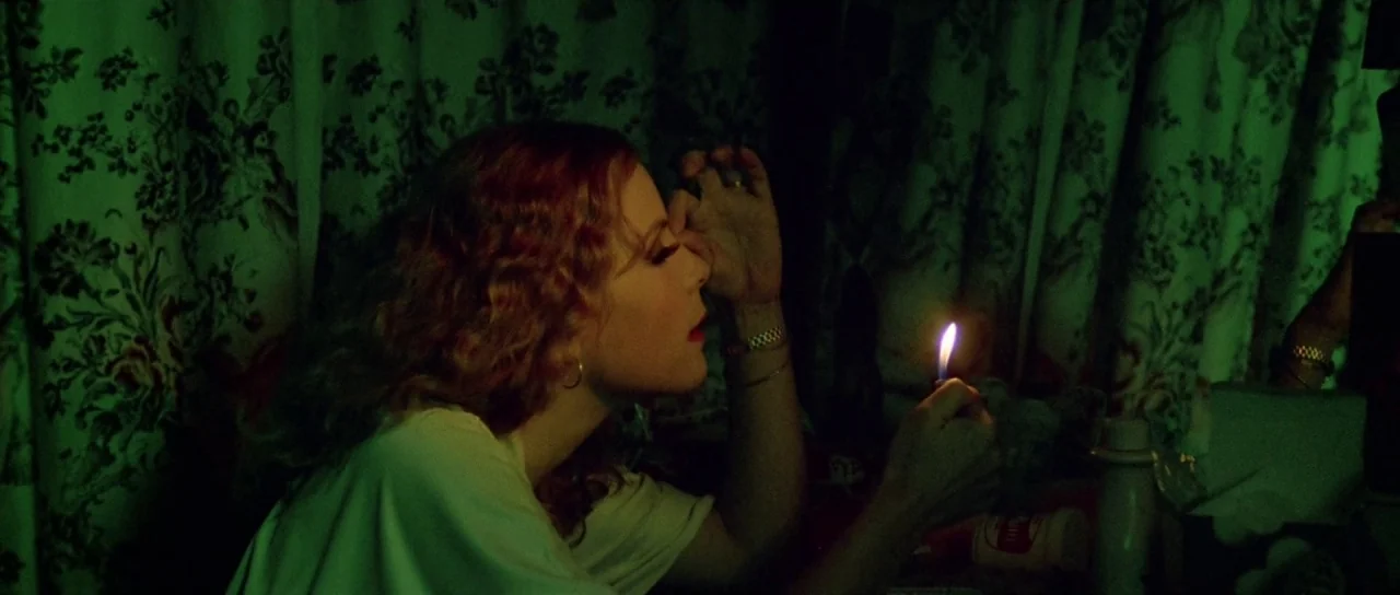 The New York Ripper (1982) movie still: closeup — A woman stares into a mirror in a dark, green-lit environment with floral curtains. She holds a…