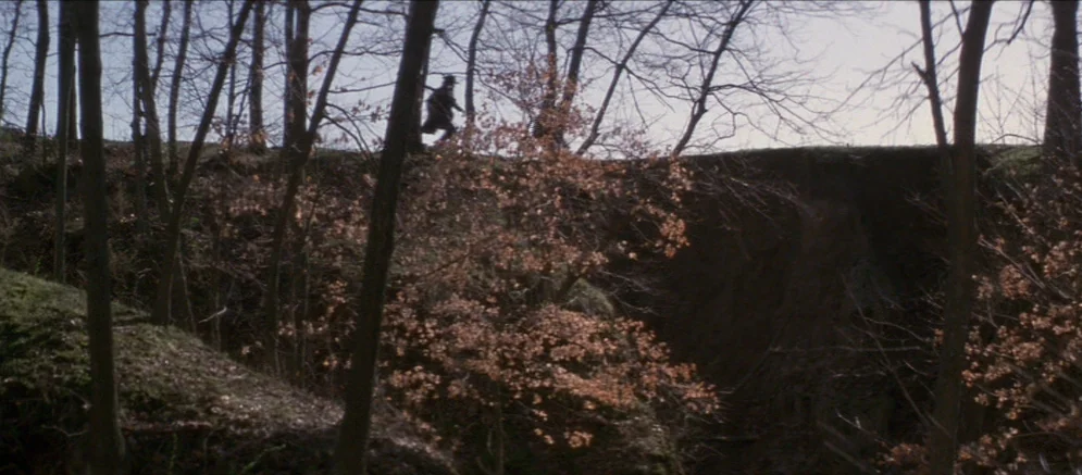 Ride With The Devil movie still: wide — A dense woodland area features a deep ravine cut into the earth. A silhouetted figure moves quickly along…