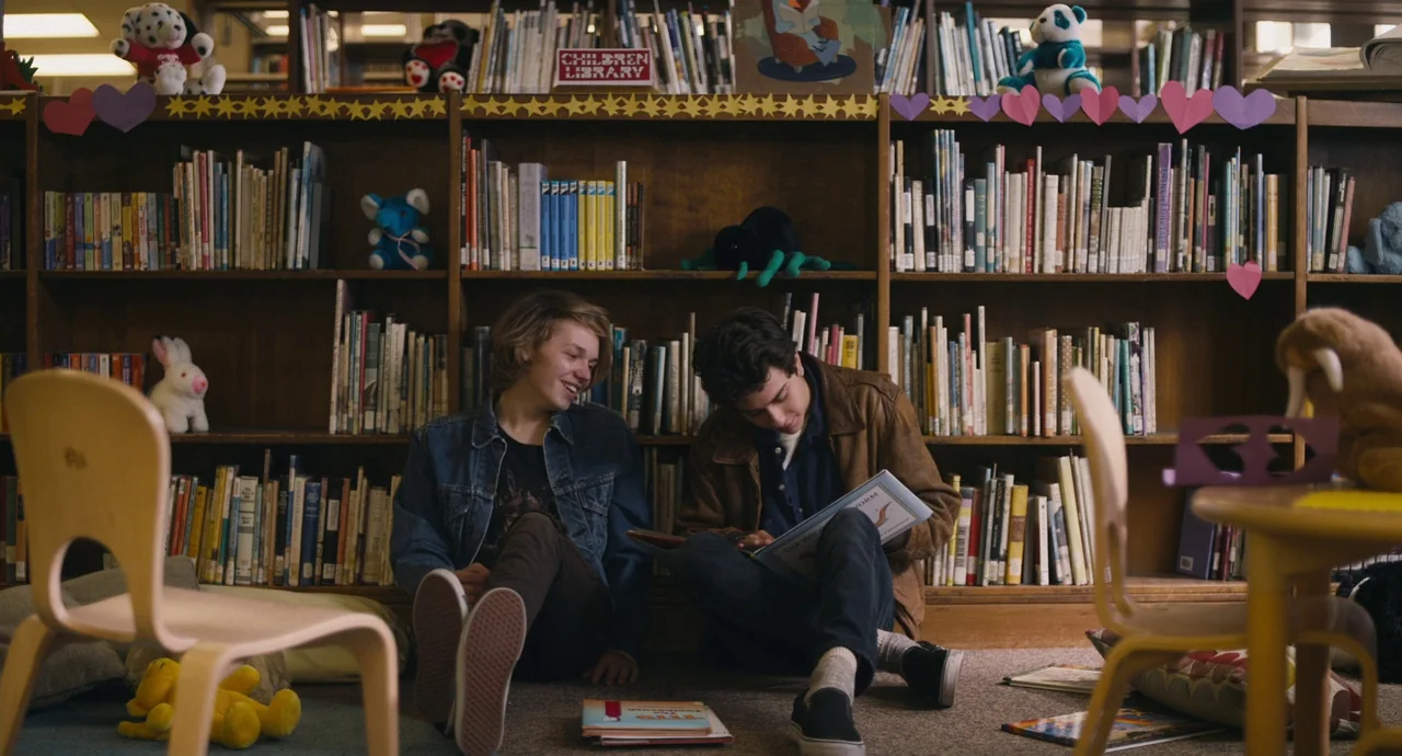 Palo Alto (2013) movie still: medium — A children's section in a library with wooden shelves filled with books and plush toys. Two teenage boys sit…