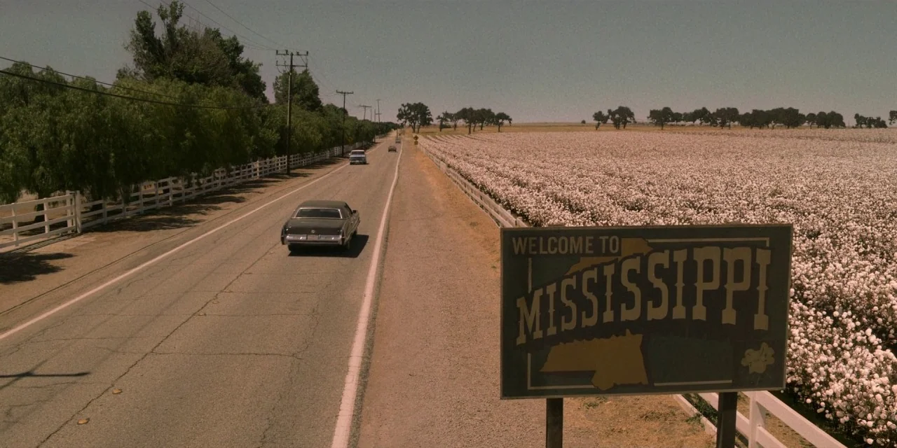 Dolemite Is My Name movie still: establishing — A long paved road stretches through a rural landscape with white picket fences. A large Welcome to…