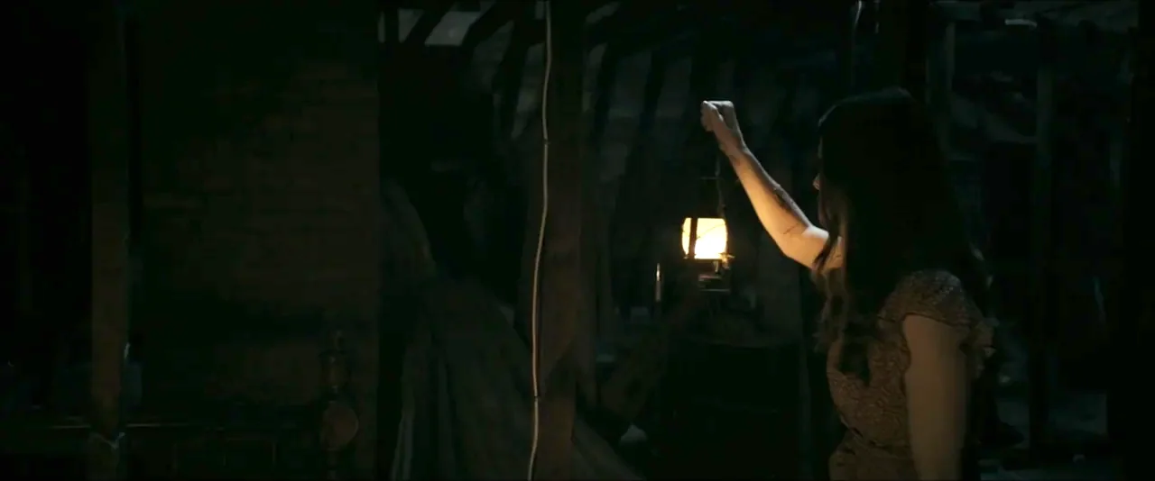 It Feeds (2025) movie still: medium — A dark attic contains wooden beams and rafters partially obscured by heavy shadows. A woman holding an…