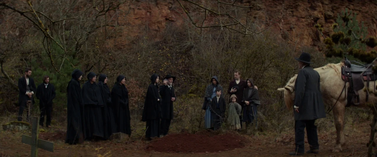 Never Grow Old (2019) movie still: wide — A rural gravesite located against a natural rock face with scattered leafless trees. A group of people…