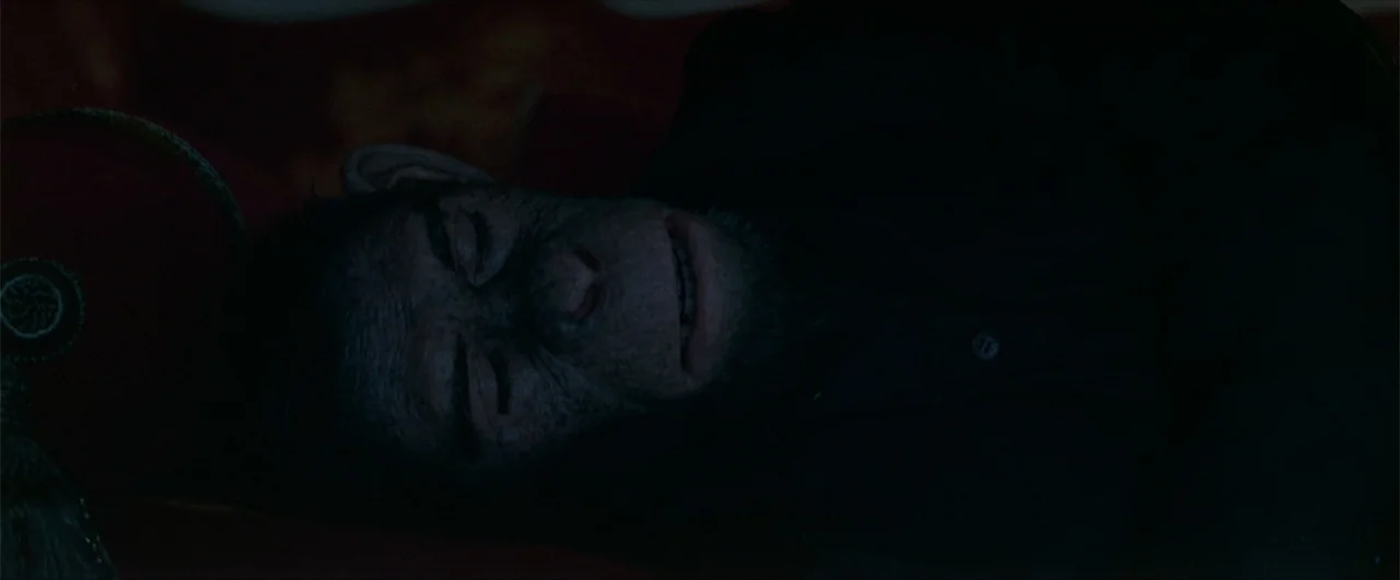 Better Man (2024) movie still: closeup — A dimly lit room features dark surfaces and a red textured cushion. A chimpanzee character lies down in the…