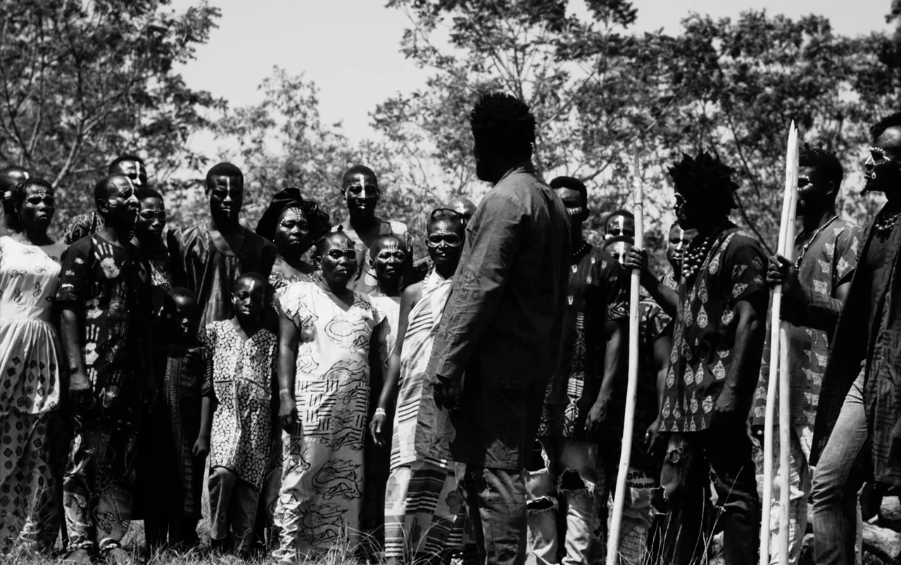 Mami Wata (2023) movie still: fullbody — A large group of people stands in a sunlit grassy field among trees. They wear patterned clothing and…