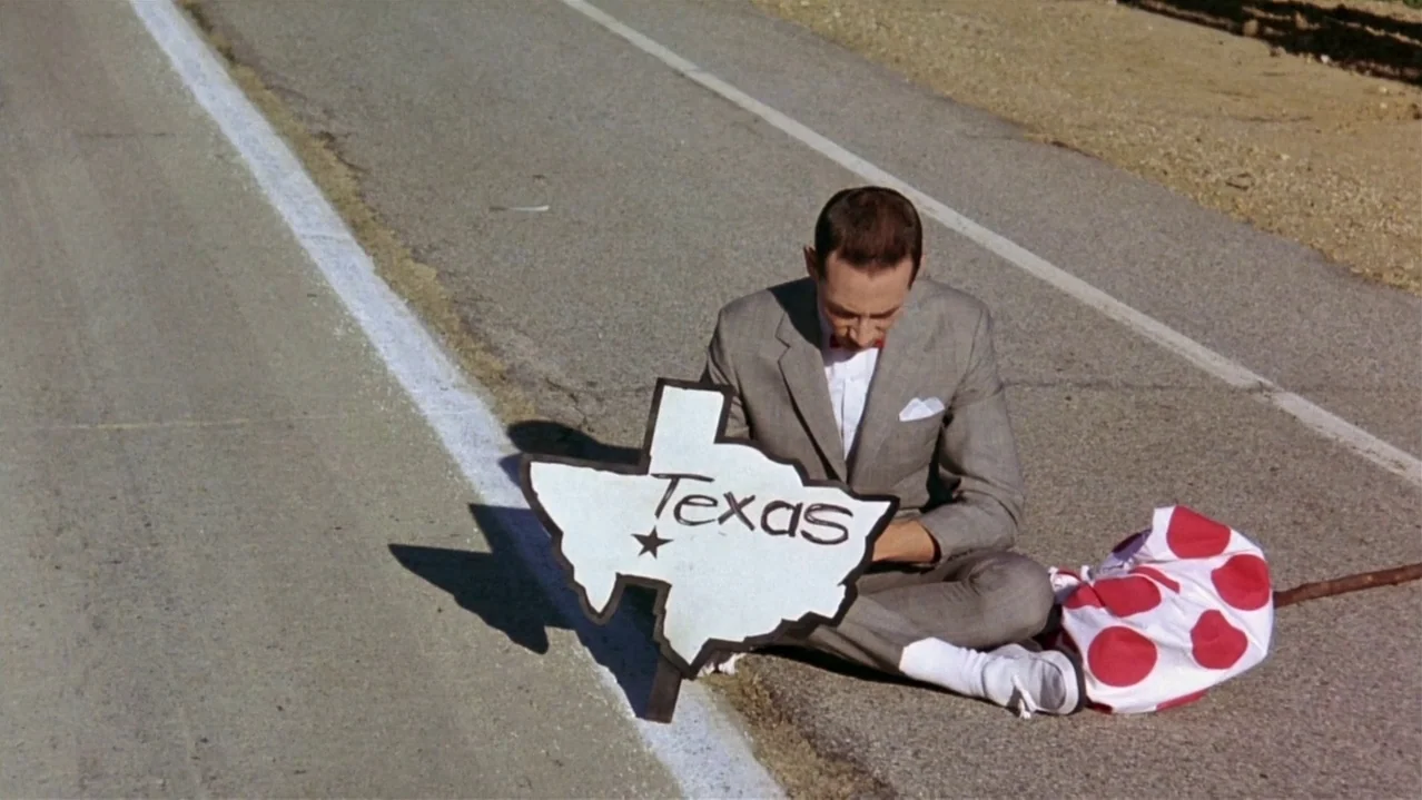 Pee-Wee’s Big Adventure (1985) movie still: medium — A man sits on the side of a paved road with a painted sign. He holds a large cutout map of…