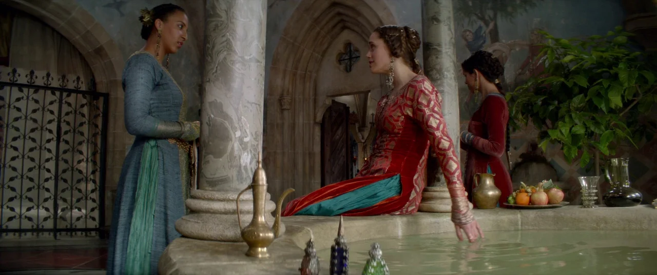 Ophelia (2018) movie still: wide — An opulent stone room with arched doorways and wall murals contains a central pool. Two women dressed in ornate…