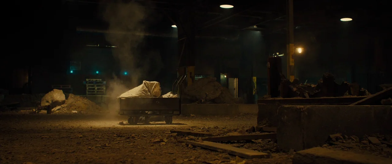 Free Fire (2016) movie still: wide — An abandoned industrial interior floor contains piles of dirt, concrete rubble, and scattered debris. A metal…