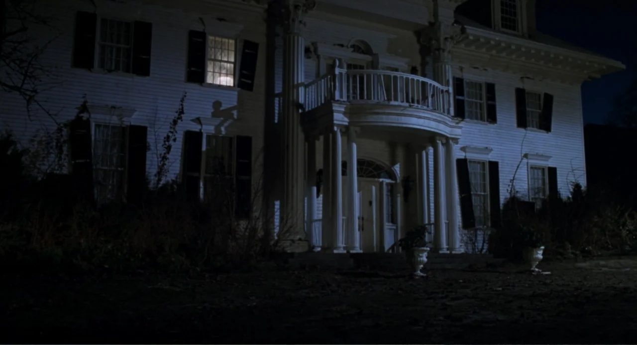 Jumanji (1995) movie still: establishing — A two-story colonial mansion features white siding and a covered porch at night. Light glows from one…