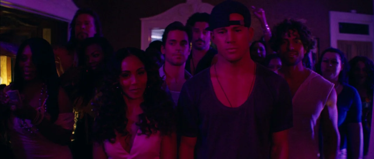 Magic Mike XXL (2015) movie still: medium — A dimly lit room is crowded with people illuminated by vibrant pink and purple lights. The main subjects…