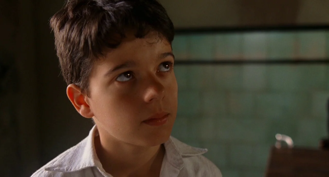 The Devils Backbone (2001) movie still: closeup — A dimly lit classroom features tiled walls of muted green in the blurred background. A young boy…