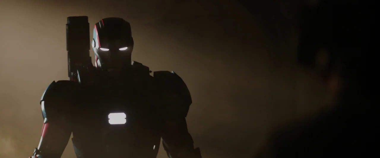 Iron Man 3 (2013) movie still: medium — A dimly lit industrial room contains an armored mechanical figure standing in the shadows. The suit's…