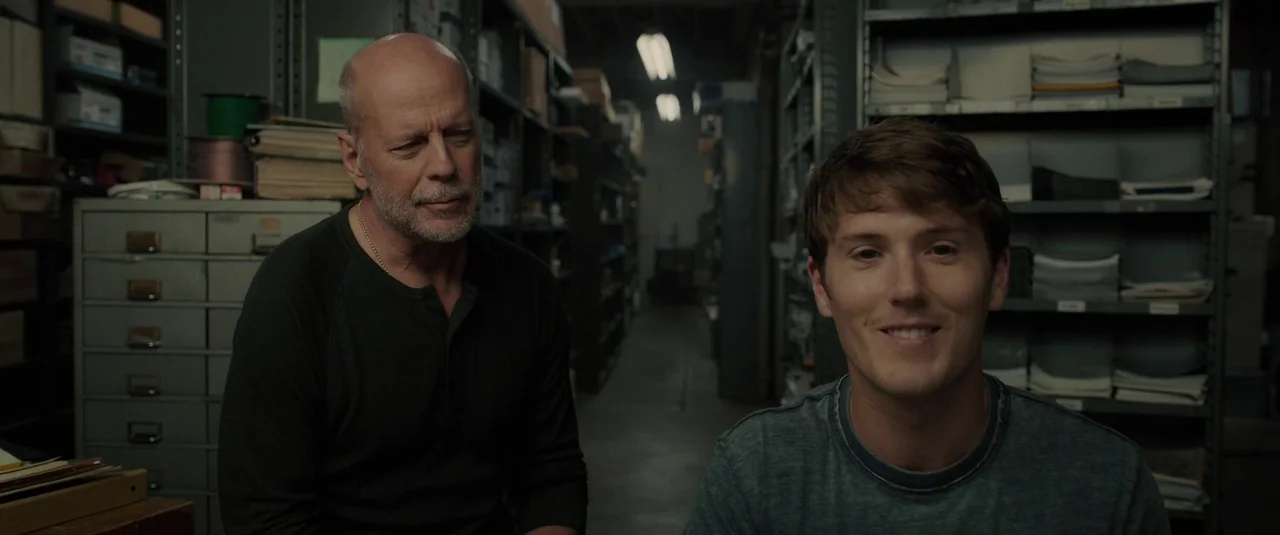 Glass (2019) movie still: medium — A cluttered industrial storage room with metal shelving units filled with documents and supplies. An older man…