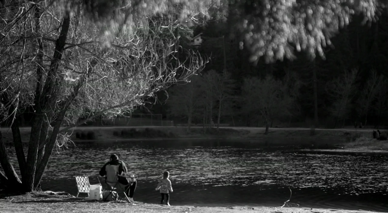 Blue Jay (2016) movie still: wide — A dark, calm lake is surrounded by bare trees in a wooded outdoor area. An adult sits in a folding chair while a…