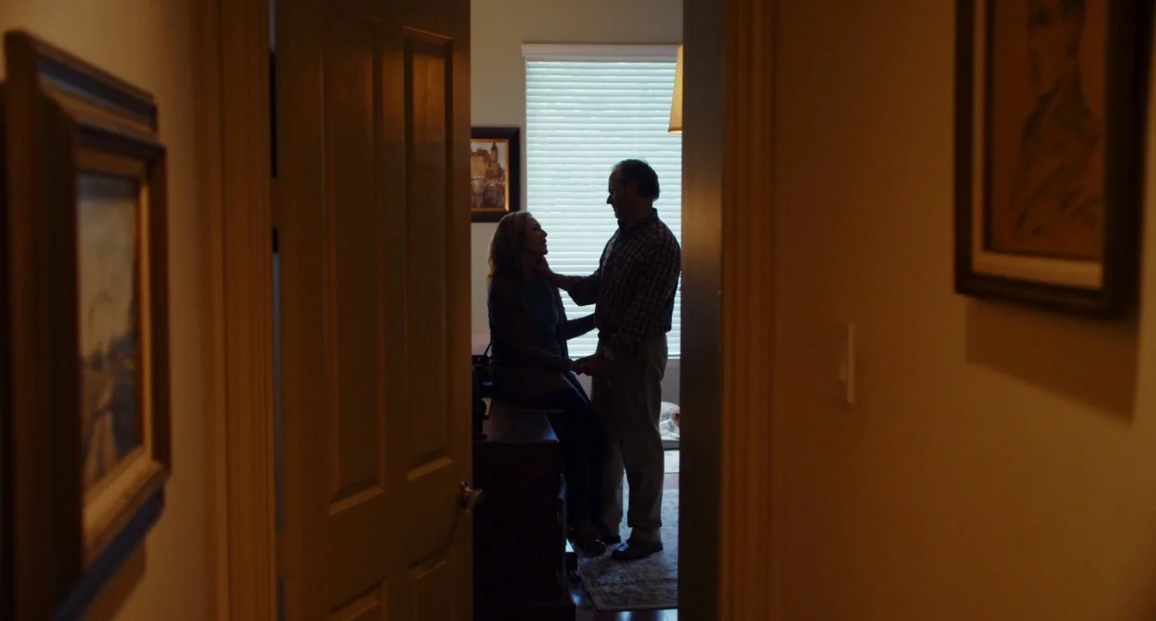Krisha (2015) movie still: wide — A view through a door frame into a room illuminated by a bright window. A man stands beside a woman who sits on…