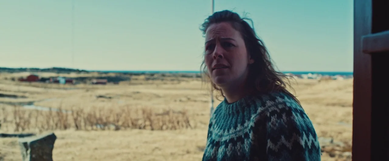 Rift (2017) movie still: medium — A vast, dry coastal field stretches toward the sea under a clear blue sky. A woman wearing a patterned wool…