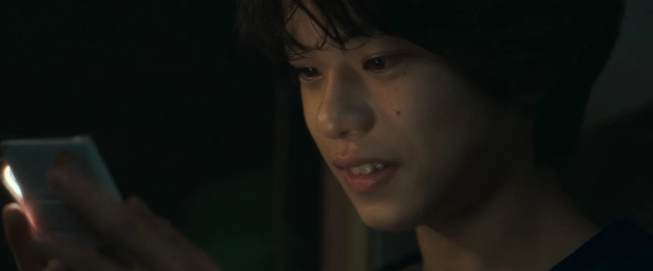 Monster (2023) movie still: closeup — A dark, indistinct indoor space at night provides the backdrop for the close-up shot. A young man stares…