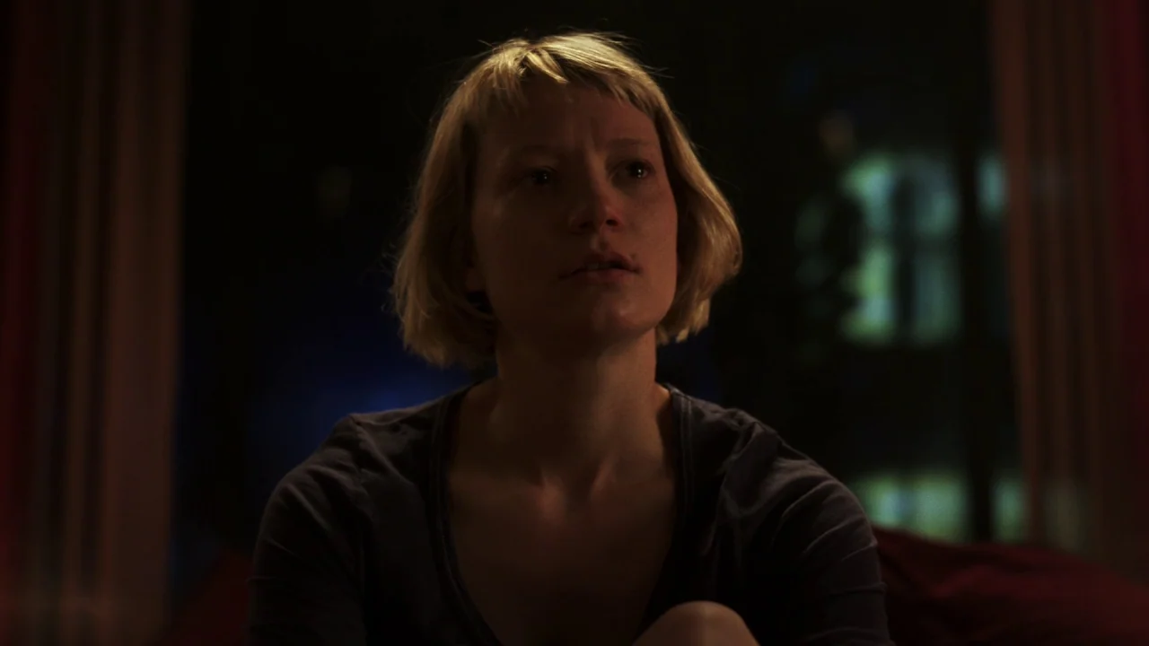 Piercing (2018) movie still: medium — A dimly lit room with dark surroundings and indistinct background shapes. A young woman with short hair stares…
