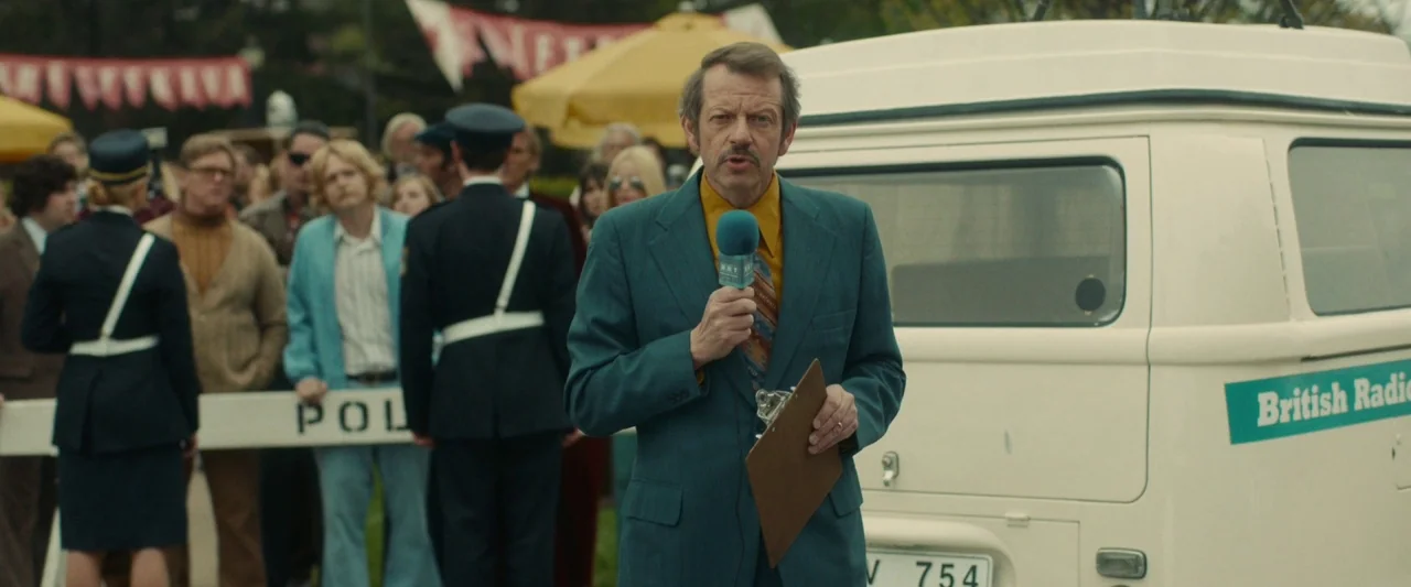 Stockholm (2019) movie still: medium — A man stands outdoors in front of a white radio van at a public event. He holds a microphone and a clipboard…