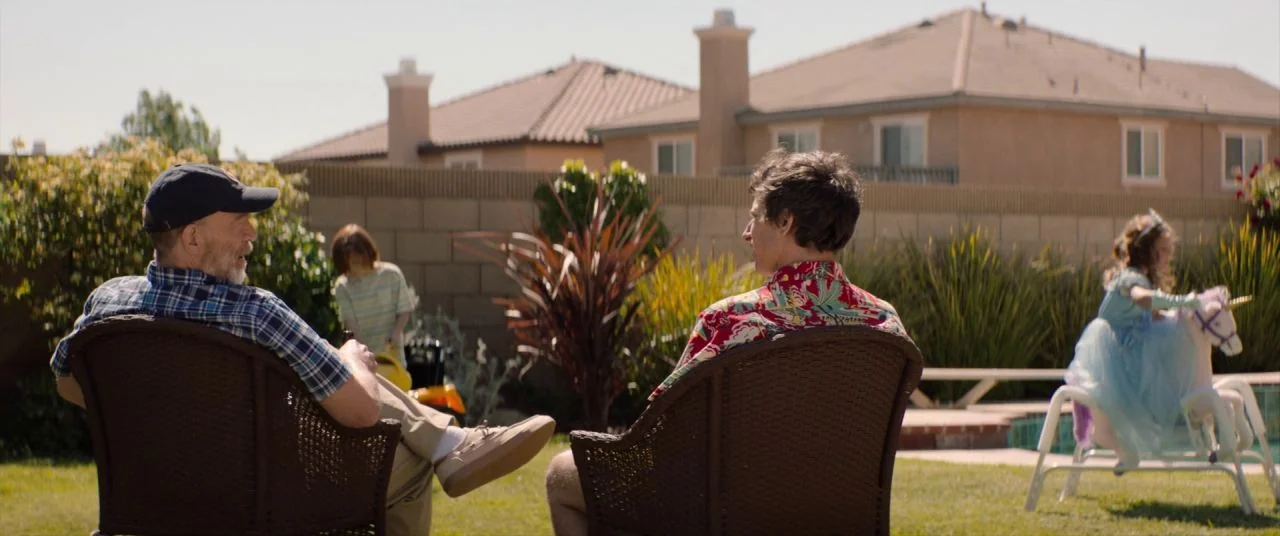 Palm Springs (2020) movie still: wide — A suburban backyard features a lawn and a cinder block wall perimeter fence. Two men sit in wicker chairs…