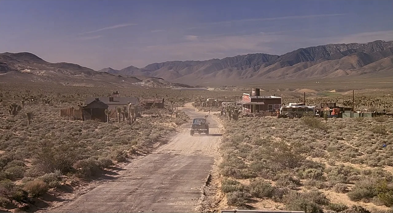 Tremors (1990) movie still: wide — An isolated desert road winds through sparse scrub brush toward distant mountain ranges. A single pickup truck…