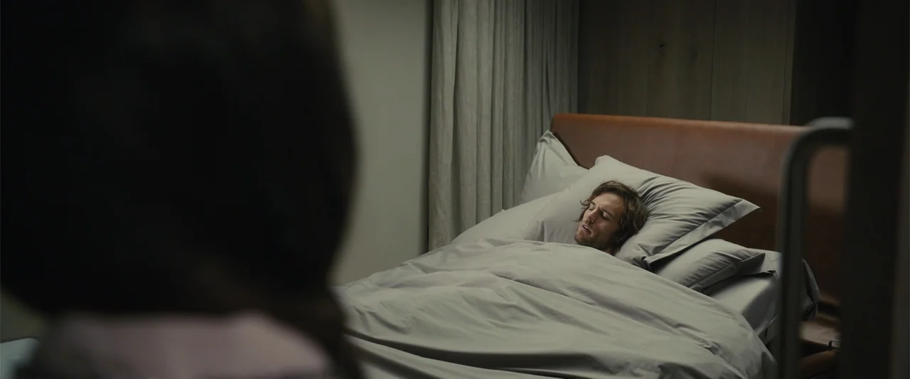 Me Before You (2016) movie still: medium — A modern bedroom with a brown leather headboard and gray bedding. A man lies in the bed with his head…