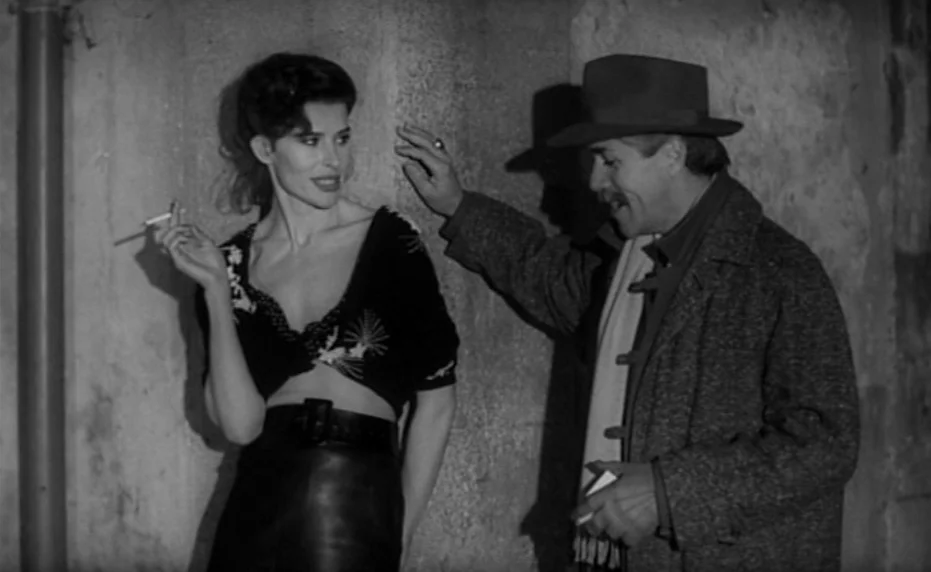 Vivement Dimanche (1983) movie still: medium — A dark outdoor space with a rough textured wall surface behind the characters. A man in a hat…