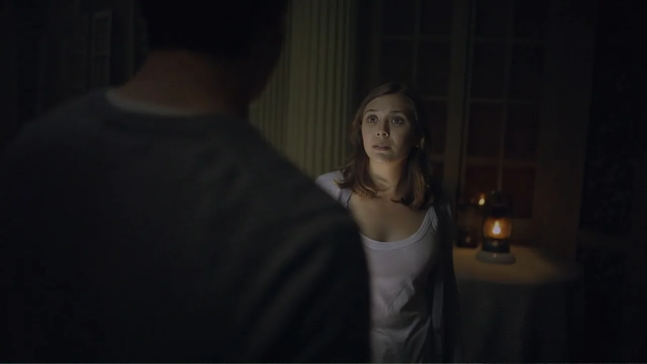 Silent House (2011) movie still: medium — A dimly lit room features a woman standing near a lit oil lamp. She faces a man whose back is turned to…