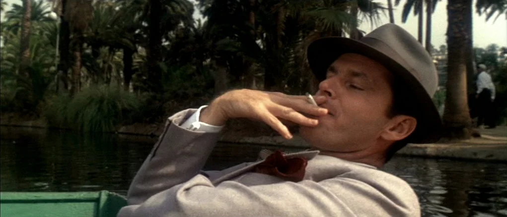 Chinatown (1974) movie still: medium — A man in a fedora relaxes in a small boat on a calm lake. Tall palm trees line the water's edge in the…