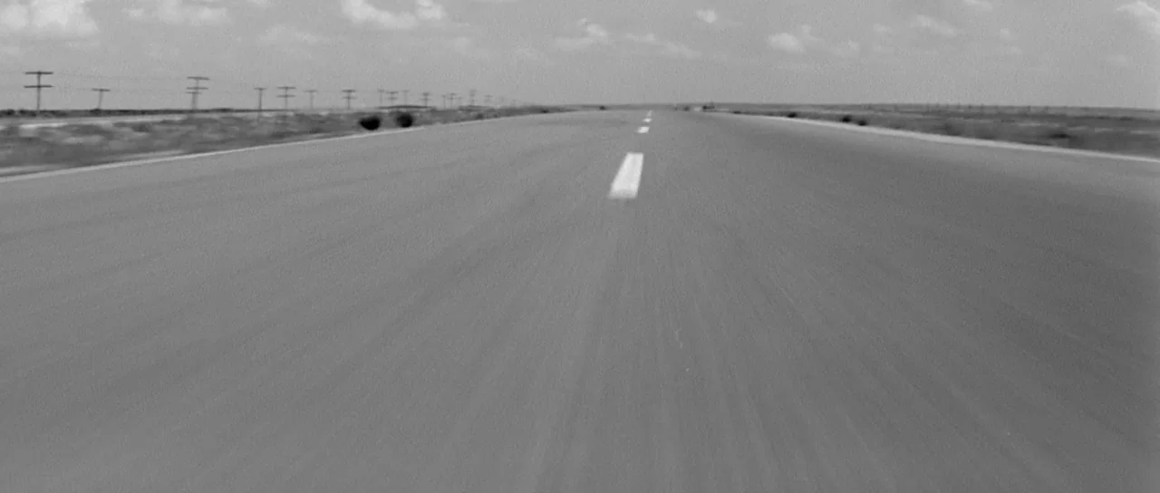 Hud (1963) movie still: establishing — A long, empty stretch of asphalt road extends toward a flat desert horizon line. Utility poles run parallel…