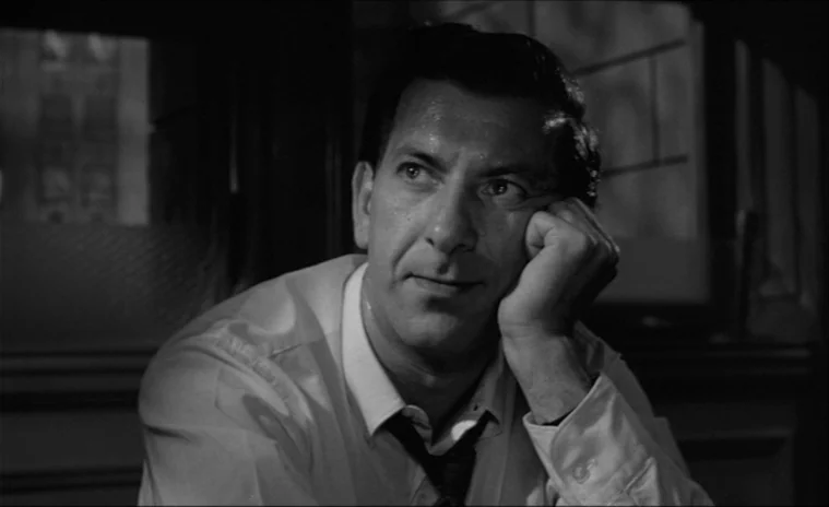 12 Angry Men (1957) movie still: medium — A dimly lit room with wood-paneled walls and a window grid in the background. A man sits at a table…