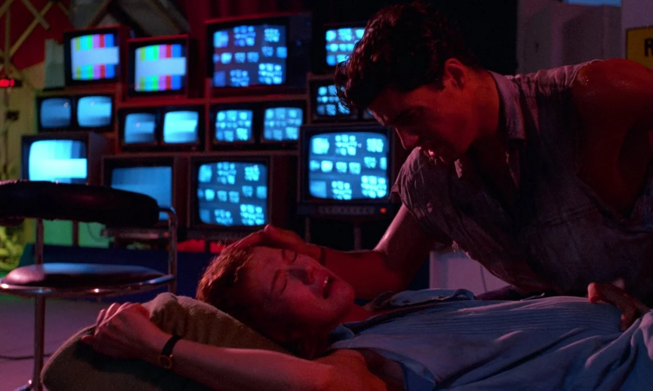 Demons 2 (1986) movie still: medium — A dark control room features a wall filled with glowing stacked television monitors. A man leans over a woman…