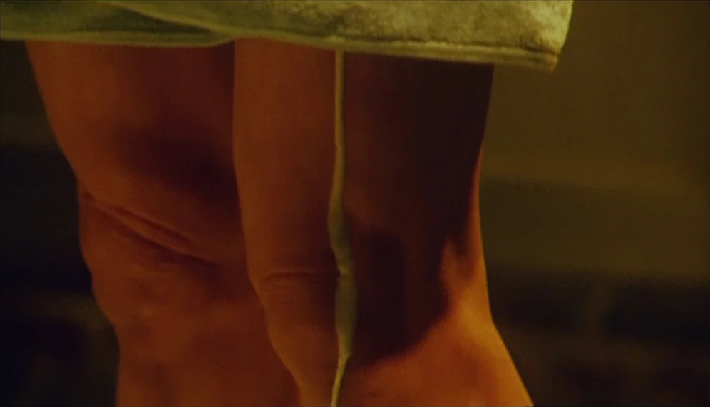 Gozu (2003) movie still: closeup — The frame shows a close-up of human legs covered by a draped bath towel. A thick vertical seam or fold of the…