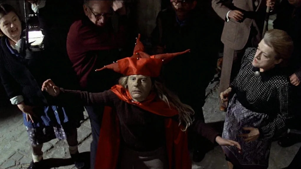 The Tenant (1976) movie still: medium — A group of people gathers on a dark, stone-paved street at night. A man wearing a bright red jester hat with…