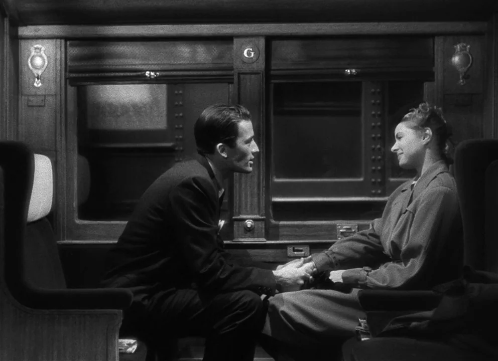 Spellbound (1945) movie still: medium — A dimly lit passenger train compartment with wood-paneled walls and a window behind them. A man and a woman…