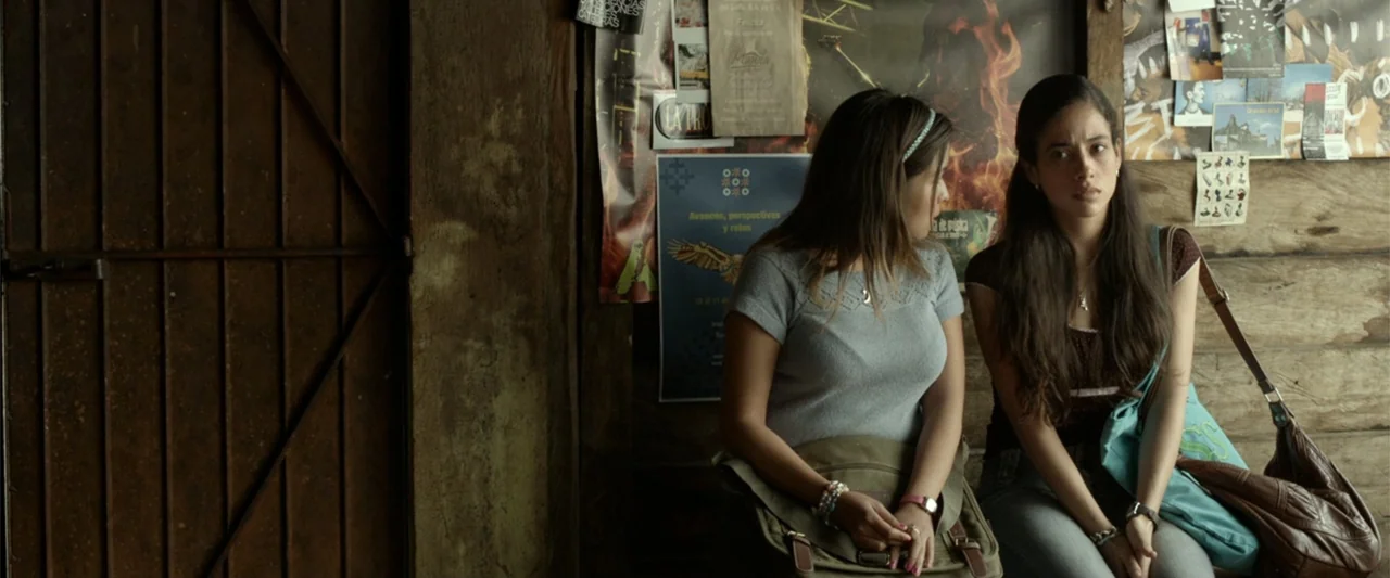 I Dream in Another Language (2017) movie still: medium — Two women sit against a dark wooden wall covered in various posted flyers indoors. They are…