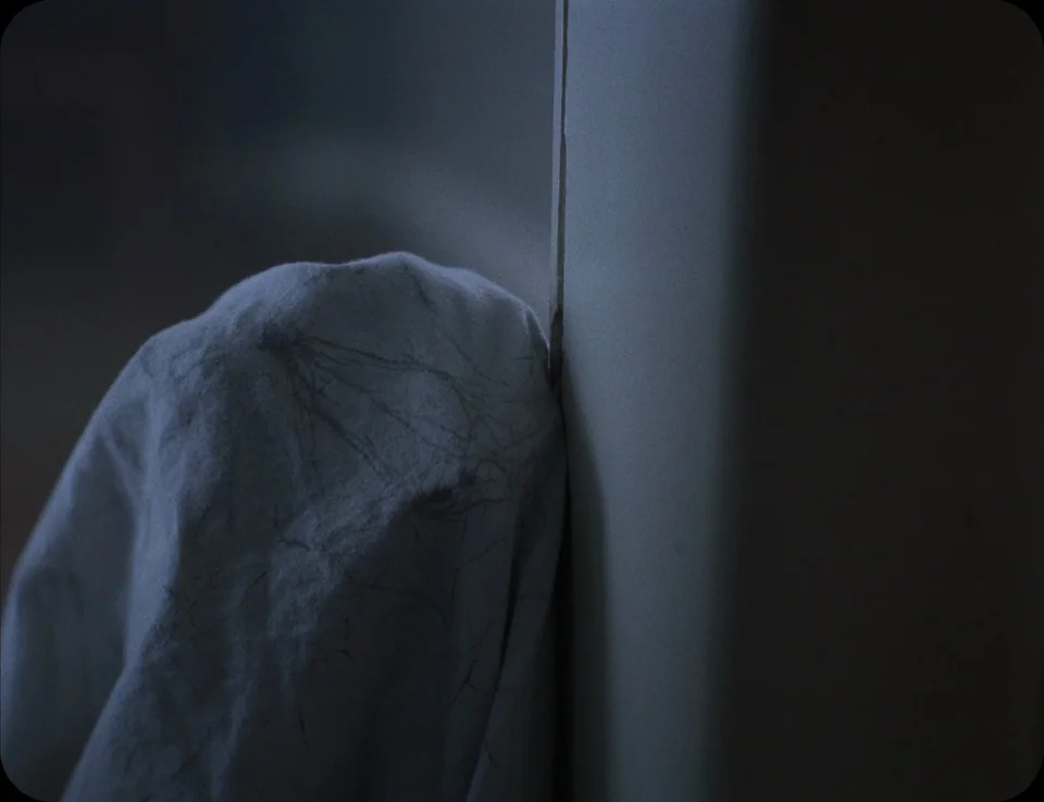 A Ghost Story (2017) movie still: closeup — A dark space containing a soft, wrinkled white textile pressed against a vertical pillar. The texture of…