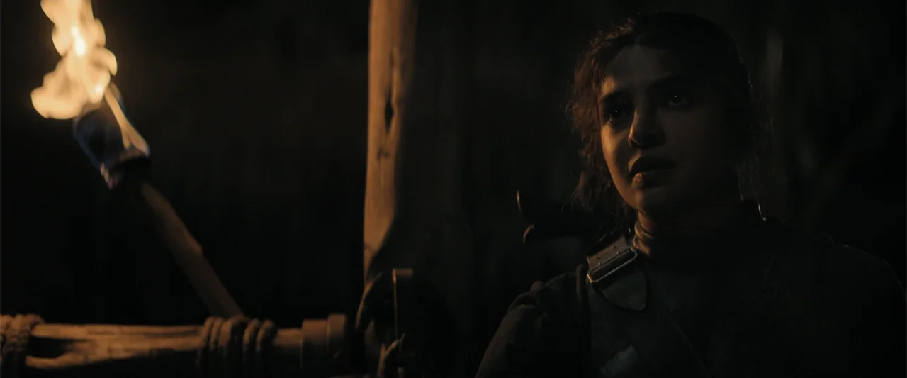 The Bluff (2026) movie still: medium — A dark, cavernous space features rugged wooden posts and low light. A woman in dark tactical gear looks out…