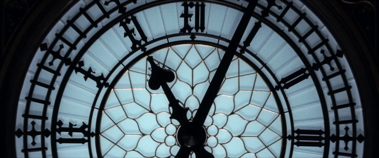 V for Vendetta (2006) movie still: closeup — A close-up view of the intricate illuminated clock face of Big Ben at night. The black metal hands are…