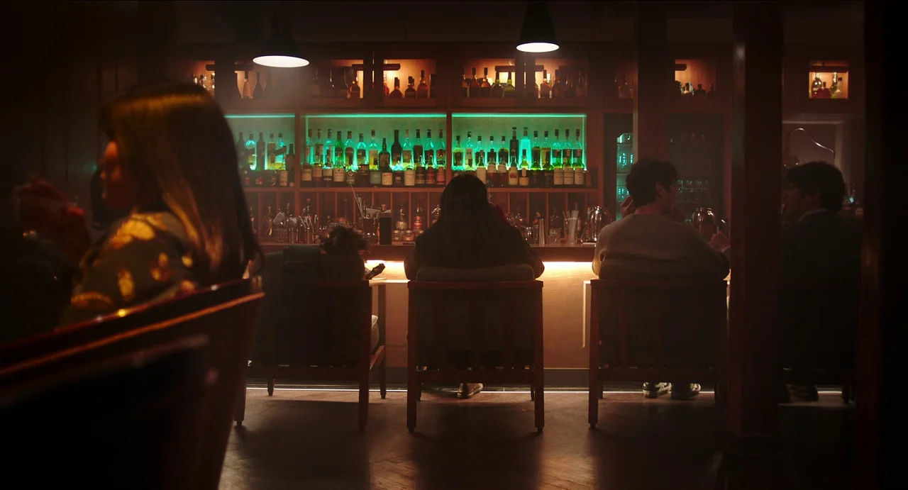 Microhabitat (2017) movie still: wide — A dimly lit lounge featuring a long wooden counter and illuminated display shelves. Multiple patrons sit on…