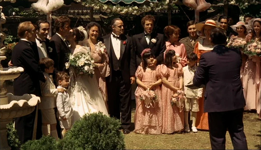 The Godfather (1972) movie still: fullbody — A group of formally dressed wedding guests gather outdoors near a decorative garden fountain. A…