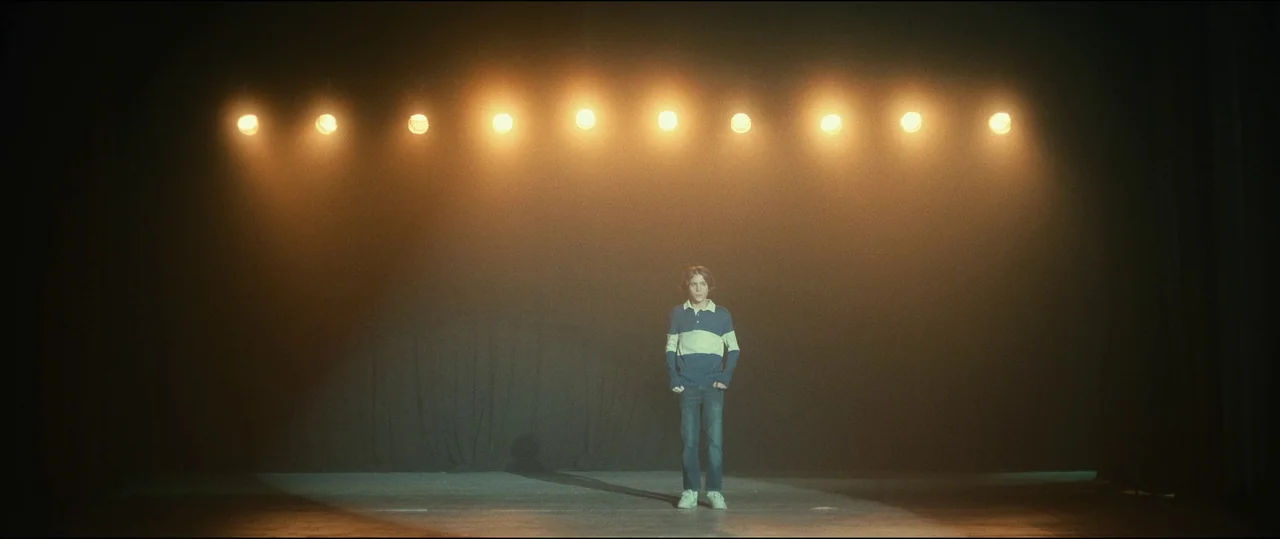 The Toxic Avenger (2023) movie still: wide — A dark empty performance area features a row of lit spotlights overhead. A single boy stands in the…