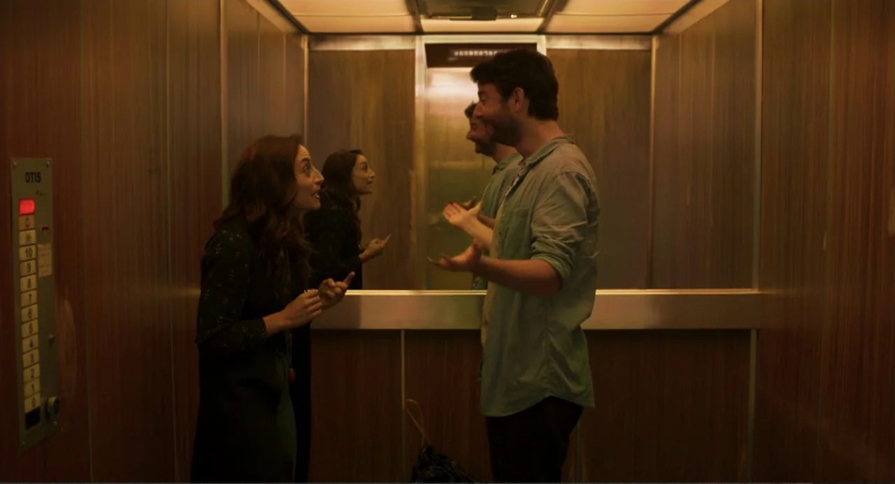 The Lift (2021) movie still: medium — A wood-paneled elevator car features a metal control panel with floor buttons. A woman and man stand face to…