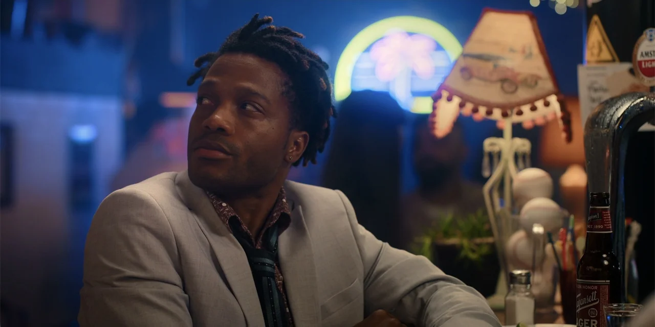 Ricky Stanicky (2024) movie still: medium — A dimly lit bar interior features a glowing neon palm sign in the background. A man sits at the counter…