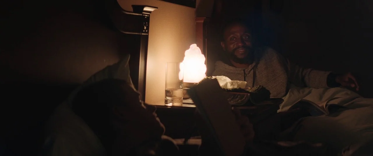 Honey Boy (2019) movie still: medium — A dark bedroom with two people resting in bed at night. One man watches his partner as they read a book by…