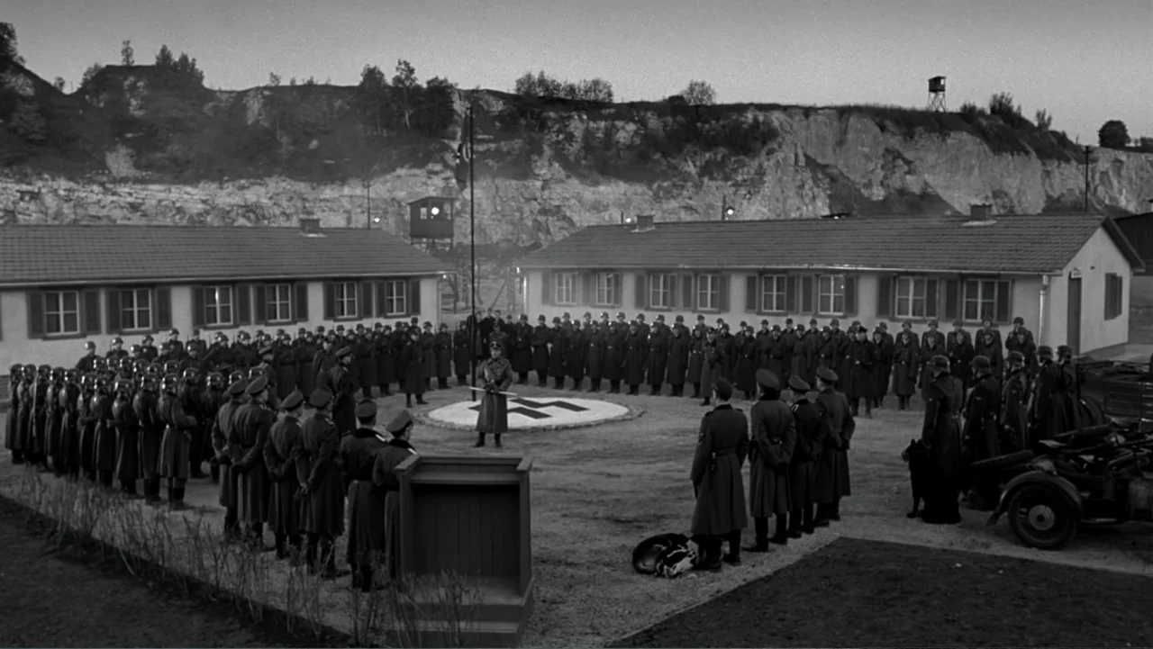 Schindler's List (1993) movie still: wide — A military camp courtyard sits at the base of a tall rocky limestone cliff. Rows of soldiers stand in a…