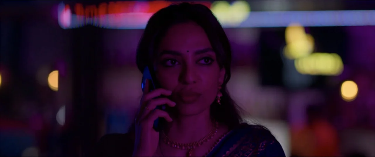 Cheekatilo (2026) movie still: closeup — A woman stands on a dark city street at night illuminated by colorful neon. She holds a mobile phone to her…