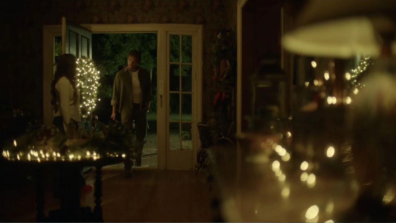 A Carpenter Christmas Romance (2024) movie still: medium — A dimly lit home entryway features floral wallpaper and a table decorated with lights. A…