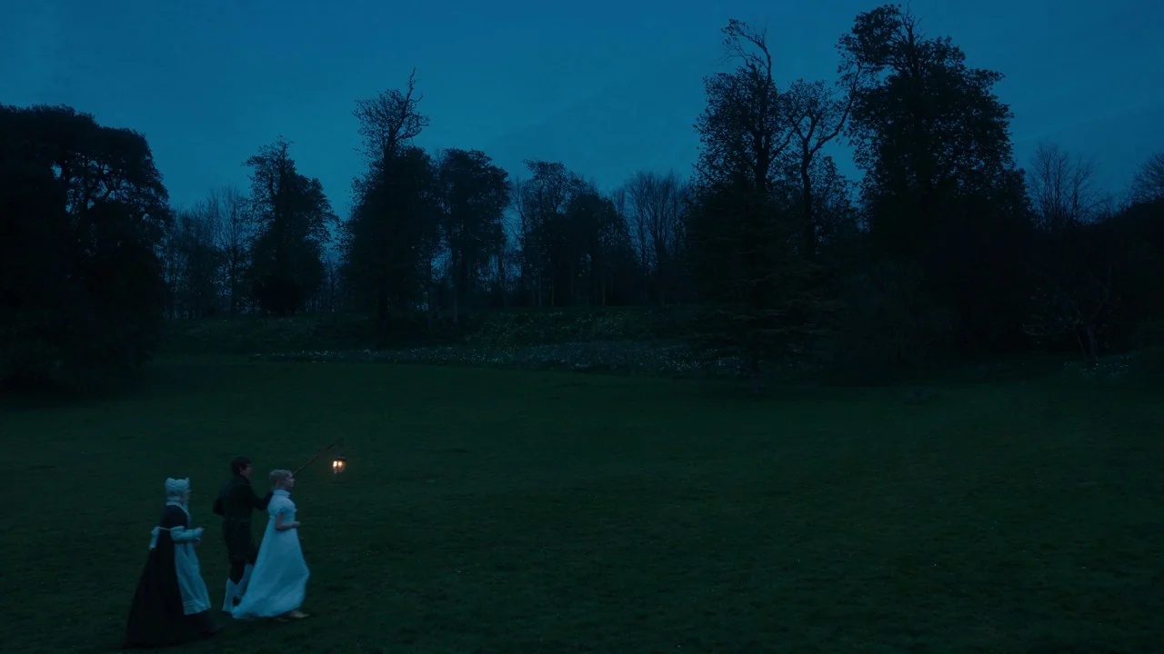 Emma. (2020) movie still: wide — A vast, dark, grassy field is framed by a dense line of silhouetted trees. Two women and a man walk through the…